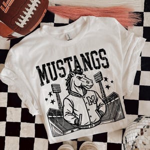 May include: White t-shirt with the word "MUSTANGS" in black block letters. A black and white illustration of a horse wearing a letterman jacket is below the word. The shirt is on a black and white checkered surface.