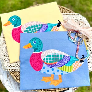 May include: Two t-shirts with a colorful duck design. One shirt is light yellow, the other is light blue. The duck has a teal head, pink chest, and a patchwork of patterns on its body. Orange beak and feet complete the design. A necklace and hair tie are also visible.