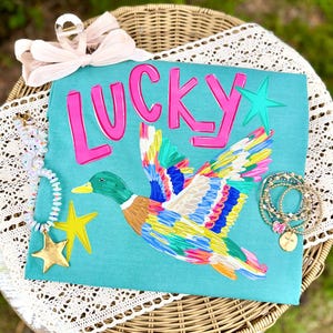 May include: A teal t-shirt featuring the word LUCKY in bold pink letters, a multi-colored mallard duck in flight, and a teal star. The shirt is accessorized with a light pink hair bow, a beaded necklace with a gold star, and stacked beaded bracelets.