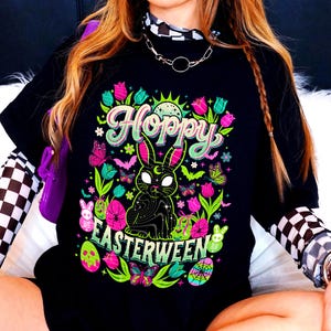 May include: Black t-shirt with a colorful graphic design. The design features the word "Hoppy" in green and pink, a black rabbit with a crescent moon, and the word "Easterween". The design also includes butterflies, flowers, and Easter eggs.