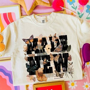 May include: Cream-colored t-shirt with the words "MADE NEW" in large black block letters. The letters are surrounded by floral and butterfly graphics in shades of brown, orange, and purple. The shirt has a classic crew neck.