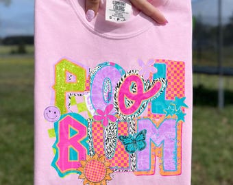 May include: A light pink sweatshirt with a colorful graphic design. The design includes the words "POOL ROOM" in various fonts and patterns, along with a smiley face, flowers, a butterfly, and stars. The sweatshirt is held up by a hand.