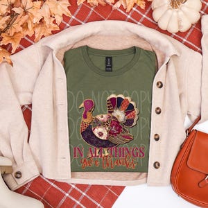 May include: An olive green t-shirt with a colorful turkey design and the text "IN ALL THINGS give thanks". The shirt is layered under a beige jacket, with a brown purse and white boots.