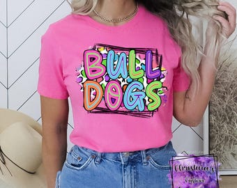 May include: A bright pink t-shirt with the word "BULLDOGS" in colorful, bubble-style letters. The text is set against a leopard print background within a black rectangular frame. The shirt is short-sleeved.