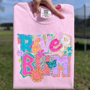 May include: A light pink sweatshirt with the words "RIVER RUM" in a colorful, whimsical design. The design includes a smiley face, flowers, stars, a butterfly, and a sunflower. The sweatshirt is made by Comfort Colors.