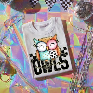 May include: White t-shirt featuring a colorful graphic of two owls with closed eyes. The owls are in shades of blue, pink, and orange, with a black and white spotted pattern. The word "OWLS" is printed in large black letters.