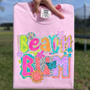 May include: A light pink Comfort Colors sweatshirt with the words "Beach Bum" in a colorful, whimsical design. The design includes a seashell, flowers, a smiley face, a butterfly, and a sunflower. The sweatshirt is held up by a hand.