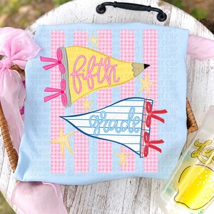 May include: A light blue t-shirt featuring a pink gingham striped background with two pennant-shaped graphics. The top yellow pencil-shaped pennant reads fifth in pink script, and the bottom white lined pennant reads grade in blue script.