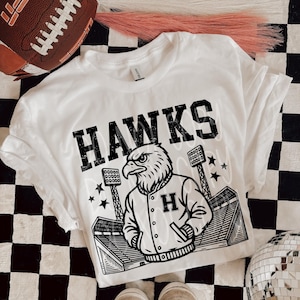 May include: White t-shirt with the word "HAWKS" in black block letters. The shirt features a graphic of an eagle wearing a letterman jacket with the letter "H". The design includes stadium lights and stars.