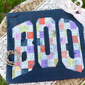 May include: A dark gray t-shirt featuring the word BOO in large block letters. The letters are filled with a multicolored checkered pattern in shades of orange, purple, yellow, and white. A stack of gold-toned beaded bracelets rests on the letter B.