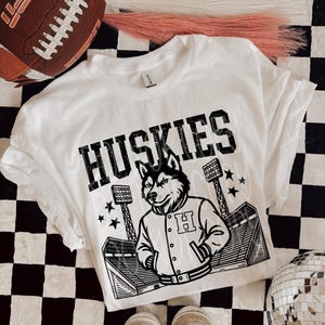 May include: White t-shirt with the word "Huskies" in bold black letters. A cartoon husky wearing a letterman jacket is centered below the word. The shirt is on a black and white checkered surface, with a football and shoes in the background.