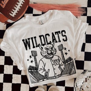 May include: White t-shirt with the word "WILDCATS" in black block letters. The shirt features a graphic of a wildcat wearing a jacket with a "W" on it, stadium lights, and stars. A brown football and sneakers are also in the image.