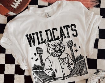 Pode incluir: Camiseta branca com a palavra "WILDCATS" em letras pretas. A camiseta apresenta um gráfico de um gato selvagem usando uma jaqueta com um "W", luzes de estádio e estrelas. Uma bola de futebol marrom e tênis também estão na imagem.