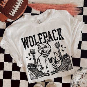 May include: White t-shirt with a black graphic that says "WOLFPACK" and features a wolf wearing a letterman jacket. The shirt also shows a stadium and stars. A brown football and shoes are also in the image.