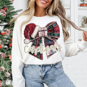 May include: White sweatshirt featuring a festive bow design with red, green, and white plaid patterns. The bow incorporates a Santa hat element. The word "Cardinals" is written in gold glitter script, creating a holiday-themed apparel.