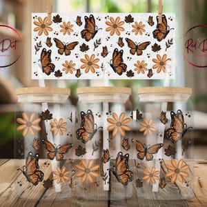 May include: Three glass jars with wooden lids and straws, decorated with butterflies, flowers, and leaves. The jars are clear, and the design is in shades of brown and orange. A sign with the same design hangs above the jars.