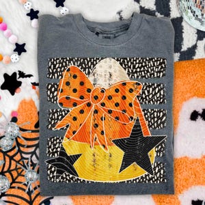 May include: A charcoal gray crewneck t-shirt featuring a graphic print of a candy corn shape adorned with a large orange polka-dot bow. The design includes two black stars, a background of horizontal black and white dashed stripes, and a distressed texture.