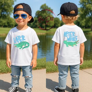 May include: White t-shirt with a green alligator graphic and the word "LATER" in blue. The back of the shirt has the words "AFTER WHILE" and a matching alligator. The child is wearing blue jeans and a black baseball cap.