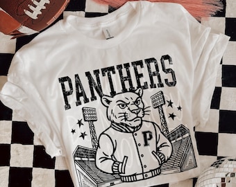 Pode incluir: Camiseta branca com a palavra "PANTHERS" em letras pretas em negrito. A camiseta apresenta um gráfico de uma pantera usando uma jaqueta com um "P", junto com um estádio e estrelas. Uma bola de futebol marrom e tênis também estão na imagem.