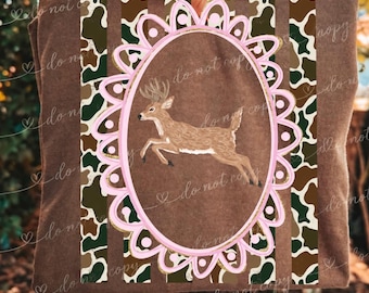 May include: A brown t-shirt featuring a central oval frame with a scalloped pink border. Inside the frame is a brown deer in a leaping pose. The background of the shirt displays vertical stripes of brown and green cow print patterns.