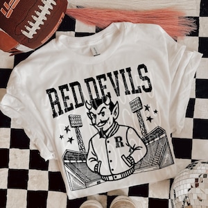 May include: White t-shirt with a graphic of a devil mascot, the words "RED DEVILS", and a stadium design. A brown football and a pair of sneakers are also visible. The shirt is on a black and white checkered surface.