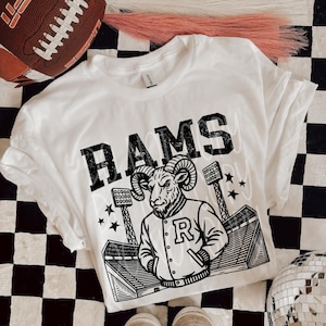 May include: White t-shirt with the word "RAMS" in large, black, block letters. The shirt features a graphic of a ram wearing a letterman jacket with the letter "R" and a stadium scene. A football and shoes are also in the image.