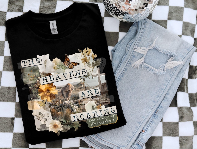 Op de afbeelding: Zwart T-shirt met een collage-ontwerp met een leeuw, bloemen en een vlinder. De tekst "THE HEAVENS ARE ROARING" staat op de voorkant. Lichtblauwe distressed jeans en een discobal zijn ook in de afbeelding.