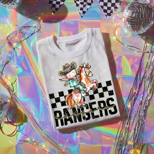 May include: White t-shirt featuring a cartoon cowboy riding a horse. The cowboy wears a hat and scarf, and the horse has a spotted pattern. The shirt has the text "Rangers" in a bold font with a checkered pattern.