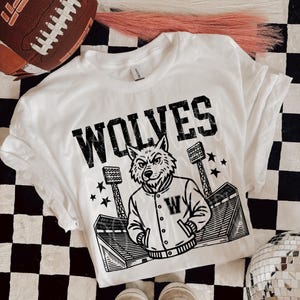 May include: White t-shirt with the word "WOLVES" in bold black letters. A cartoon wolf wearing a letterman jacket is below the word. The shirt is on a black and white checkered surface.