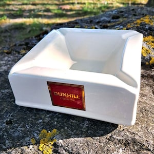 May include: A square, white ceramic ashtray with a recessed interior. The ashtray features a rectangular red and gold Dunhill Cigarettes logo on the front. The ashtray is placed on a textured, grey rock surface with yellow lichen.