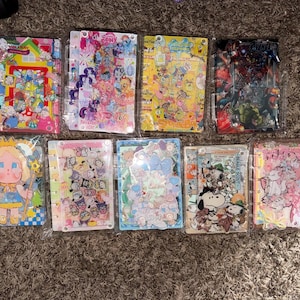 May include: A collection of colorful sticker sheets and notebooks featuring various cartoon characters and themes. The designs include My Little Pony, Marvel superheroes, and other whimsical illustrations. The items are packaged in clear plastic.
