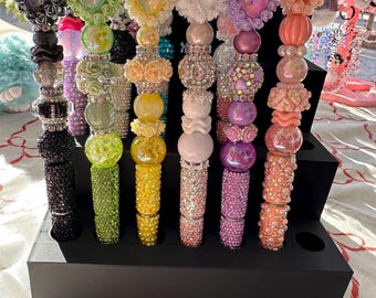 May include: A display of colorful, decorative pens with various embellishments. The pens feature sparkling rhinestones, beads, and ornate toppers in shades of black, green, yellow, white, purple, and orange. They are arranged in a black pen holder.