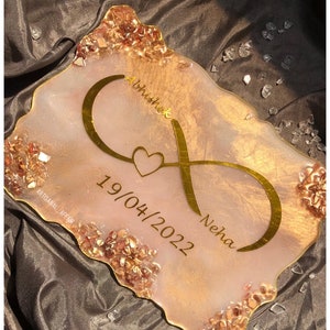 Peut inclure: Un plateau en résine rose et or avec un symbole d'infini et les noms "Abhishek" et "Neha" inscrits. La date "19/04/2022" est également inscrite sur le plateau. Le plateau est décoré de petites paillettes dorées et roses.