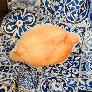 May include: A light brown, plush pillow shaped like a crescent roll. The pillow is sitting on a blue and white patterned fabric.