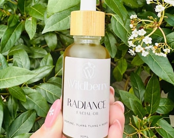 May include: A glass bottle of Wildberry Radiance Facial Oil with a bamboo lid and a white dropper. The label is white with black text that reads "Wildberry Radiance Facial Oil Jasmine, Ylang Ylang + Neroli 30ML".