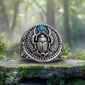 May include: A silver-toned ring featuring a central scarab beetle motif with outstretched wings. The design includes a small, round, bright blue stone at the top and a tiny, round, deep red stone at the bottom, set against a textured background.