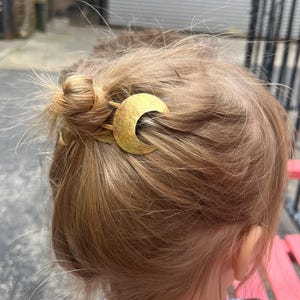 May include: A gold-colored crescent moon hair clip is placed in a bun of light brown hair. The hair clip is textured and has a hammered appearance. The image is a close-up of the back of a person's head.