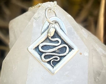 May include: A silver pendant necklace featuring a snake design inside a diamond-shaped frame. The pendant is on a silver chain and is resting on a clear crystal.