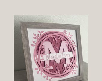 May include: A framed papercraft with a pink and white layered design of the letter M surrounded by a wreath of leaves. The name Madelyn is written on a banner below the letter.
