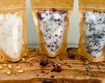 May include: Three brown paper pouches filled with bath salts. The pouches are on a wooden cutting board. The bath salts are visible through the pouches and include lavender, rose petals, and a white salt.