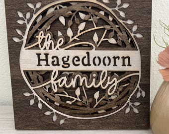 May include: A wooden sign with a decorative, laser-cut design of leaves and branches surrounding the text "the Hagedoorn family".