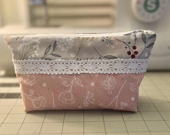 May include: A pink and gray fabric pouch with a white lace trim and a floral pattern. The pouch has a zipper closure.