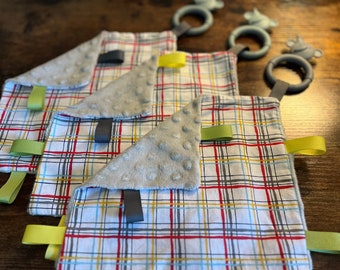 May include: Three baby blankets with a plaid pattern in shades of red, blue, yellow, and white. Each blanket has a gray, textured, triangular flap with a loop attached. The loops are connected to a set of three gray and white teething rings.