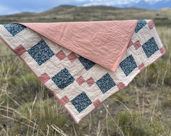 May include: A pink and white quilt with a floral pattern. The quilt is folded in half and hanging from a fence.