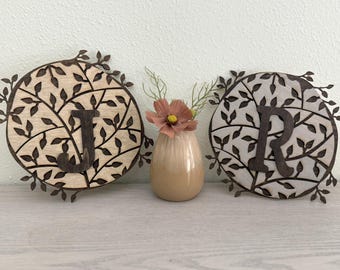 May include: Two wooden wall decor pieces with a leafy design and the letters J and R cut out. The pieces are on a light wood surface with a small vase of pink flowers in the center.