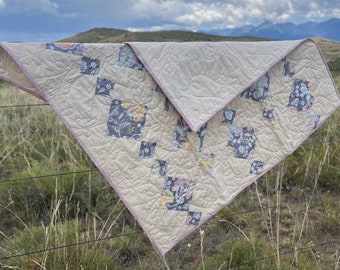 May include: A beige and gray patchwork quilt with a floral pattern. The quilt is hanging on a fence with a mountain range in the background.
