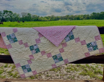 May include: A pink and cream patchwork quilt with a floral pattern. The quilt is hanging on a wooden fence with a green field and trees in the background.