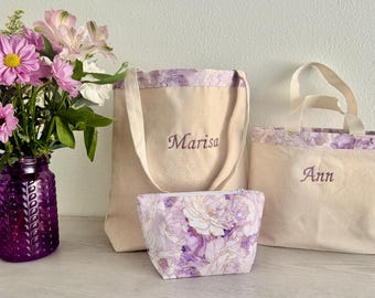May include: A still life featuring a purple glass vase with flowers, and three canvas bags. The bags are beige with purple floral trim and embroidered names. A small cosmetic bag with a floral print is also present.