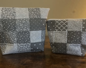 May include: Two gray and white patchwork zippered pouches. The larger pouch is on the left and the smaller pouch is on the right. Both pouches have a variety of white patterns on a gray background.