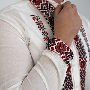 Puede incluir: Una camisa blanca de manga larga con detalles bordados en rojo y negro en el cuello y los puños. El bordado presenta patrones geométricos y motivos florales.